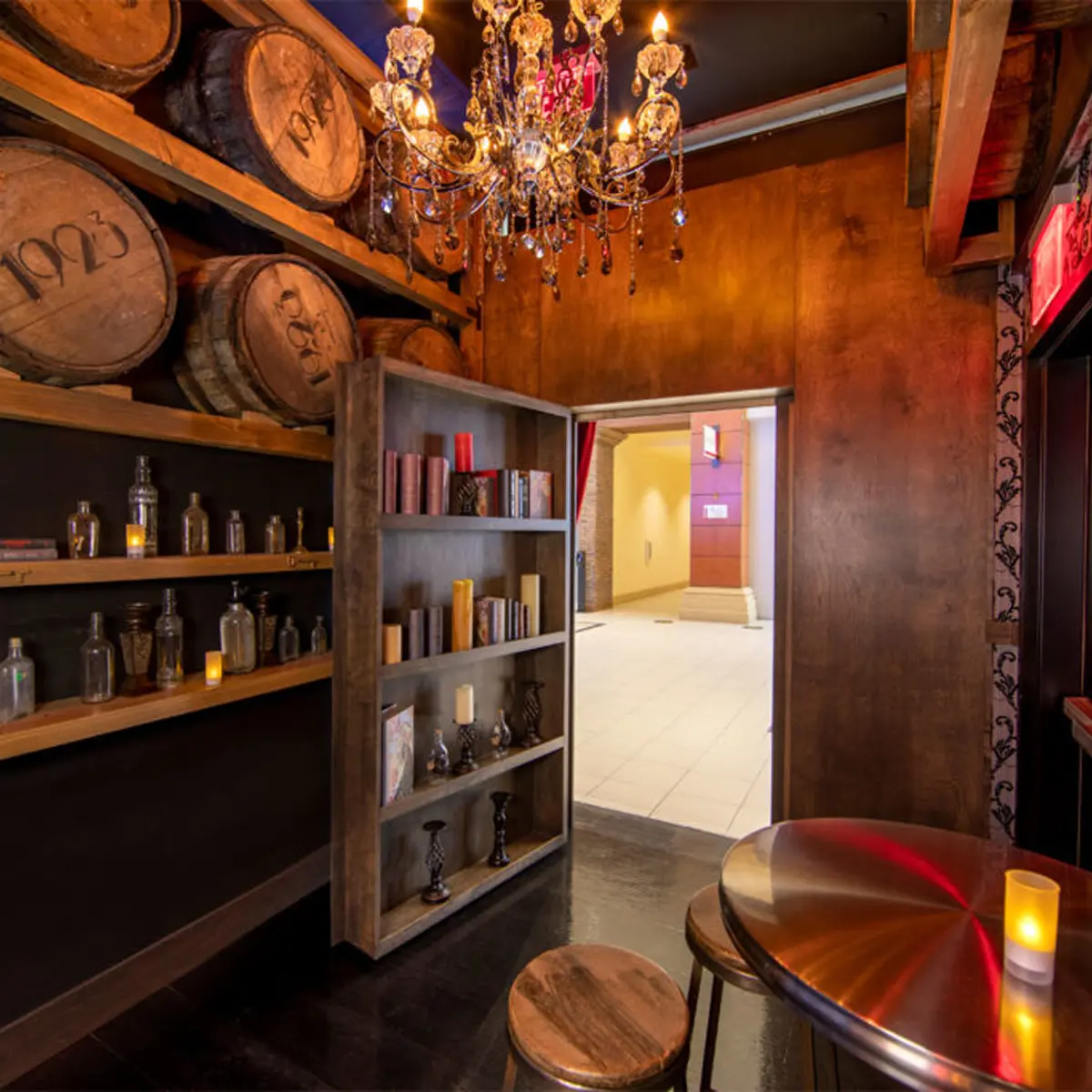 A dimly lit speakeasy-style room with stacked wooden barrels, a bookshelf secret door opening to a bright hallway, an ornate chandelier, and a small table with stools.