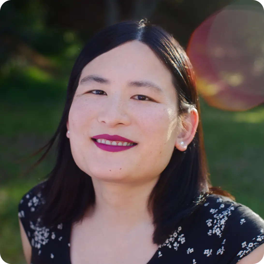 Smiling East Asian woman with dark hair, black floral top, and pearl earrings, outdoors with lens flare.