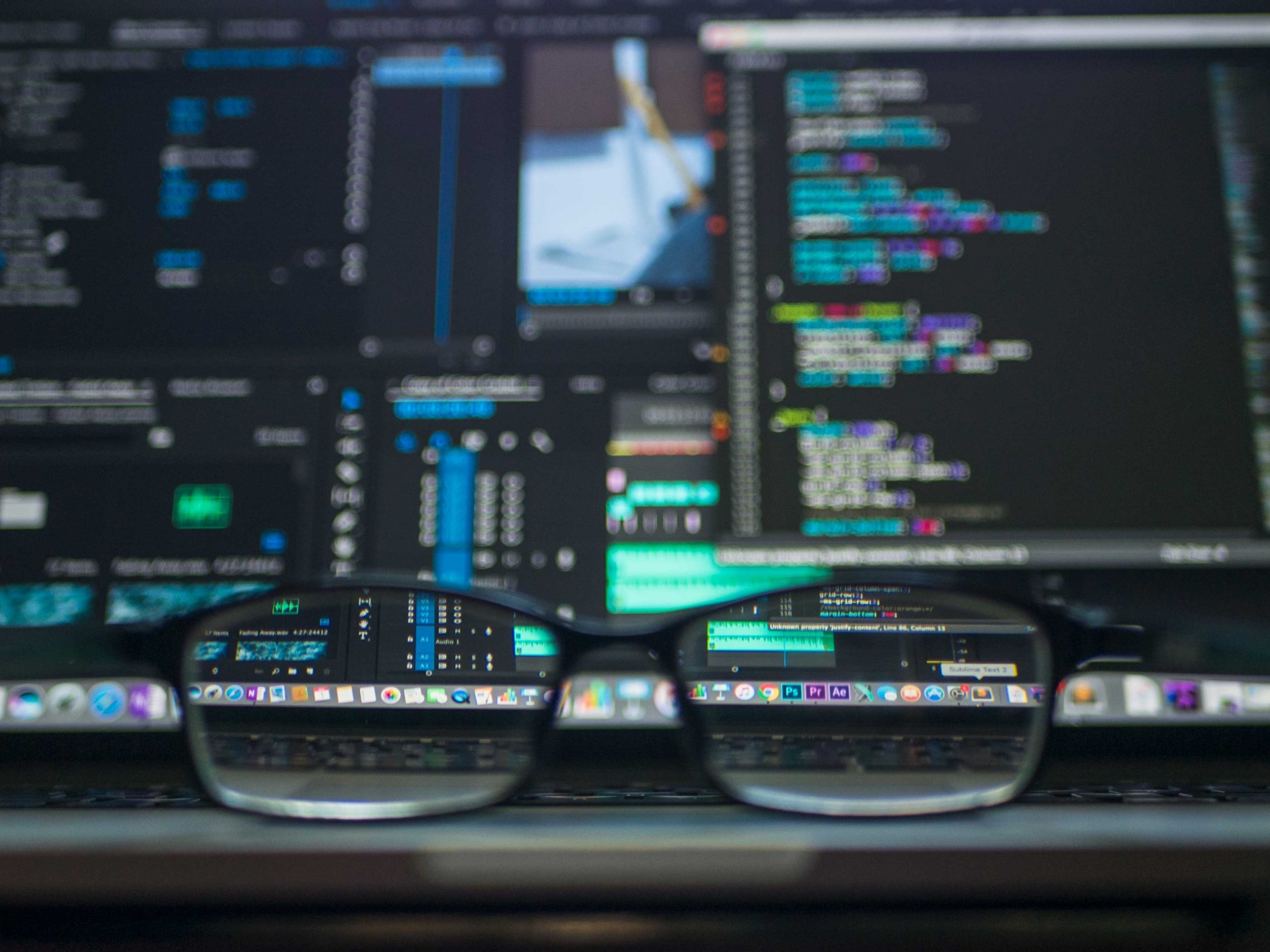 Black eyeglasses on a table with two monitors.